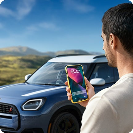 Person standing beside a MINI Countryman S ALL4, holding a smartphone and looking at the screen. 